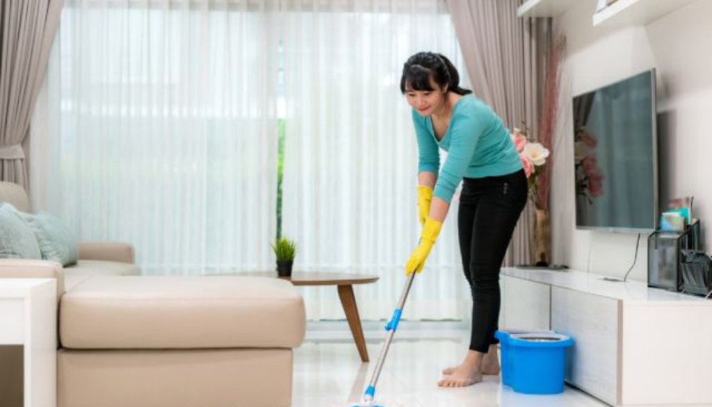Woman mopping the floor in a bright living room with yellow gloves and a blue cleaning bucket near the TV stand.