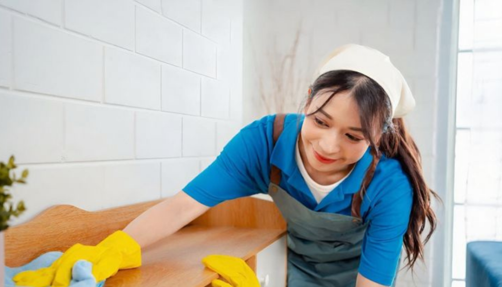 Professional house cleaner wearing gloves wiping a wooden surface in a bright room, smiling while cleaning.