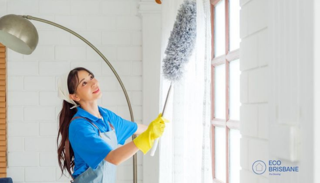 Professional cleaner dusting high window curtains with a long duster in a bright home while wearing gloves and a cleaning uniform.